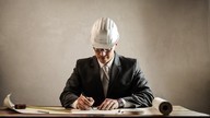 Man in hard hat and suit makes notes to plans.